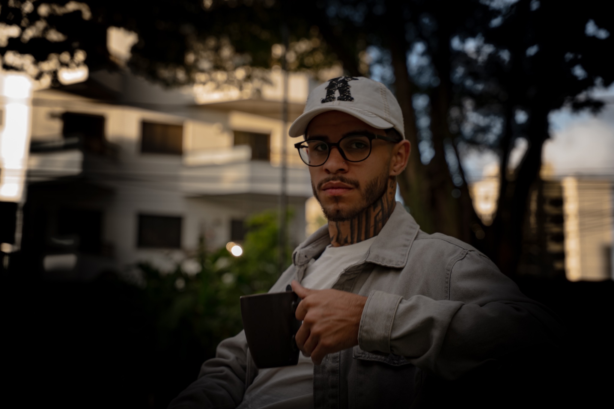Retrato de Luyd Almeida ao ar livre, de boné branco e óculos, segurando uma caneca